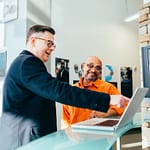 Ein Mann im Anzug zeigt einem anderen Mann in orangener Arbeitskleidung etwas begeistert auf einem Laptop. Beide sind glücklich.