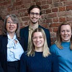 3 Frauen und 1 Mann vor einer Backsteinwand im Treppenhaus mit Blick in die Kamera und lächelnd